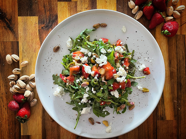 Strawberry and arugula salad on a white plate with strawberries and pistachios scattered around it on a wood board